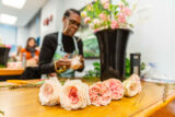a person in a light blue apron fluffs out flowers, with a bucket of flowers on a worktable and in the foreground are several fluffy, pink flowers