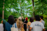 a person with red hair points up to a branch of a tree in front of a group of people