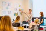 a person in a striped shirt points to landscape design drawings on a whiteboard and looks at a person with a dark blue shirt
