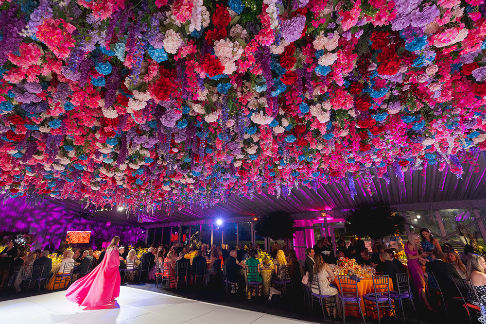 A person in a bright pink ballgown walks across a white dance floor, with pink, blue, and white flowers cascading from overhead and guests eating dinner at the edges of the space