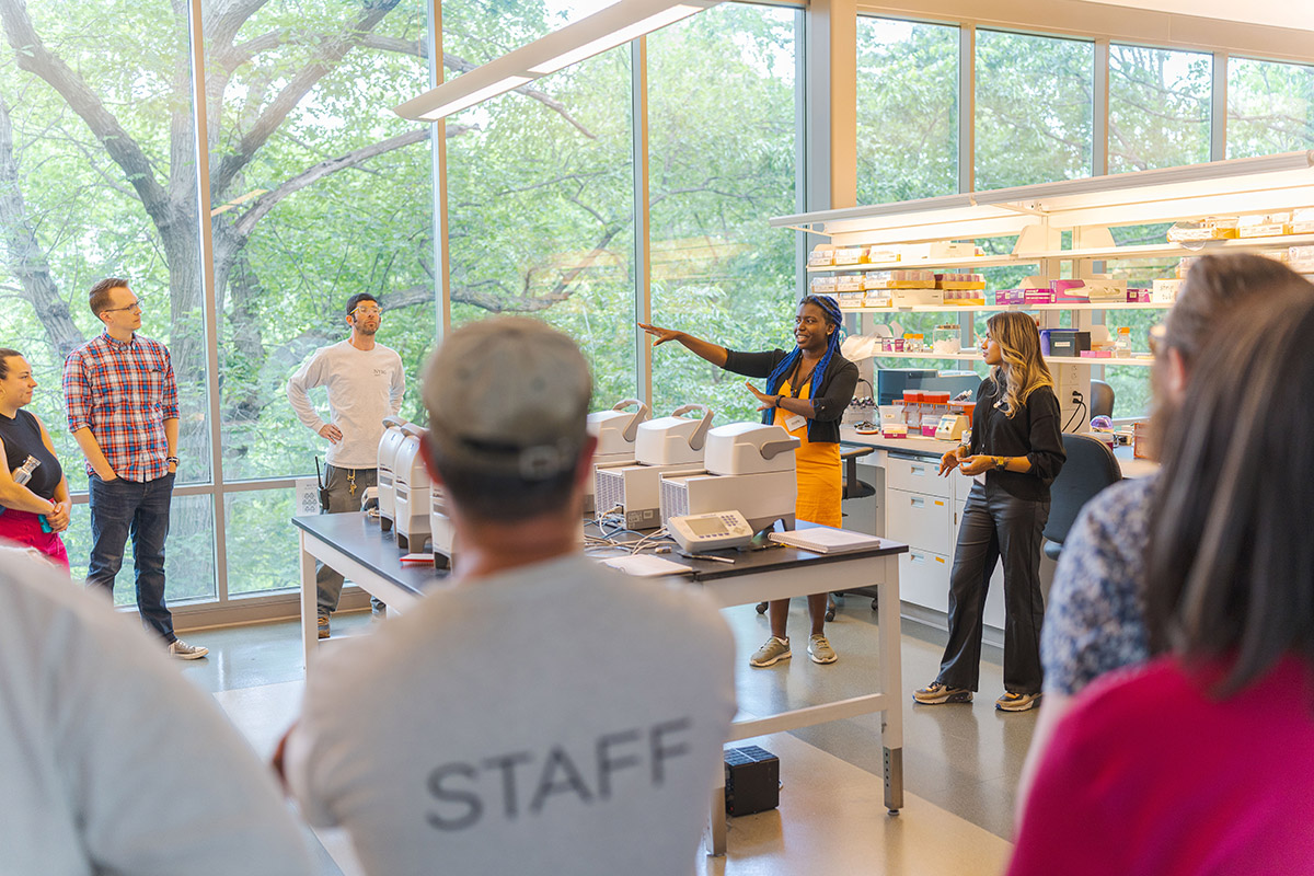 A group of people stands around a table in a brightly lit laboratory, listening to a tour guide