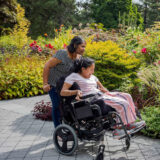 A person in a dark dress pushes another person in pink clothing, sitting in a wheelchair, as they explore a green garden space