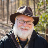 A person in a brown brimmed hat, sporting a white beard, poses for a photo in a forestal setting