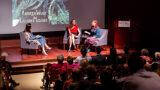 A group of people speaks in front of a crowd in an enclosed lecture hall