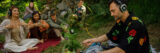 a person with headphones on sits in a wooded area with equipment in front of them connecting to the plants in the area