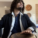 A person in a black shirt and black jacket performs on hand drums