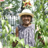 A person in a gray dress and straw hat poses for a photo among flowers and foliage