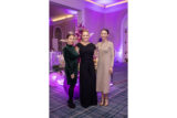 Three people in black and brown dresses pose for a photo in a holiday party setting