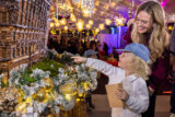 A parent and child observe a holiday scene featuring a miniature building made from dried plant parts