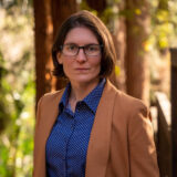 A person in a blue shirt and tan blazer poses for a photo in a forest at sunset