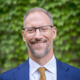 A person in a yellow tie, white shirt, and blue blazer poses outdoors for a photo