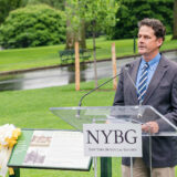 A person in a gray suit and blue shirt stands at a podium labeled "NYBG" during a rainy day outdoors