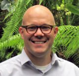 A person in a gray collared shirt and round glasses poses for a photo in a tropical location