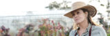 A person in a blue shirt, gardening apron, and straw hat looks on from an outdoor garden