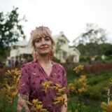 A person in a purple shirt poses in a classical garden for a photo.