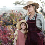 An adult and child in gardening clothes and straw hats are photographed in a garden of red flowers