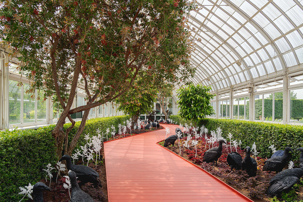 A red wooden walkway winds through a conservatory corridor of frosted glass, with small trees, shrubs, flowers, and sculptured vultures on either side