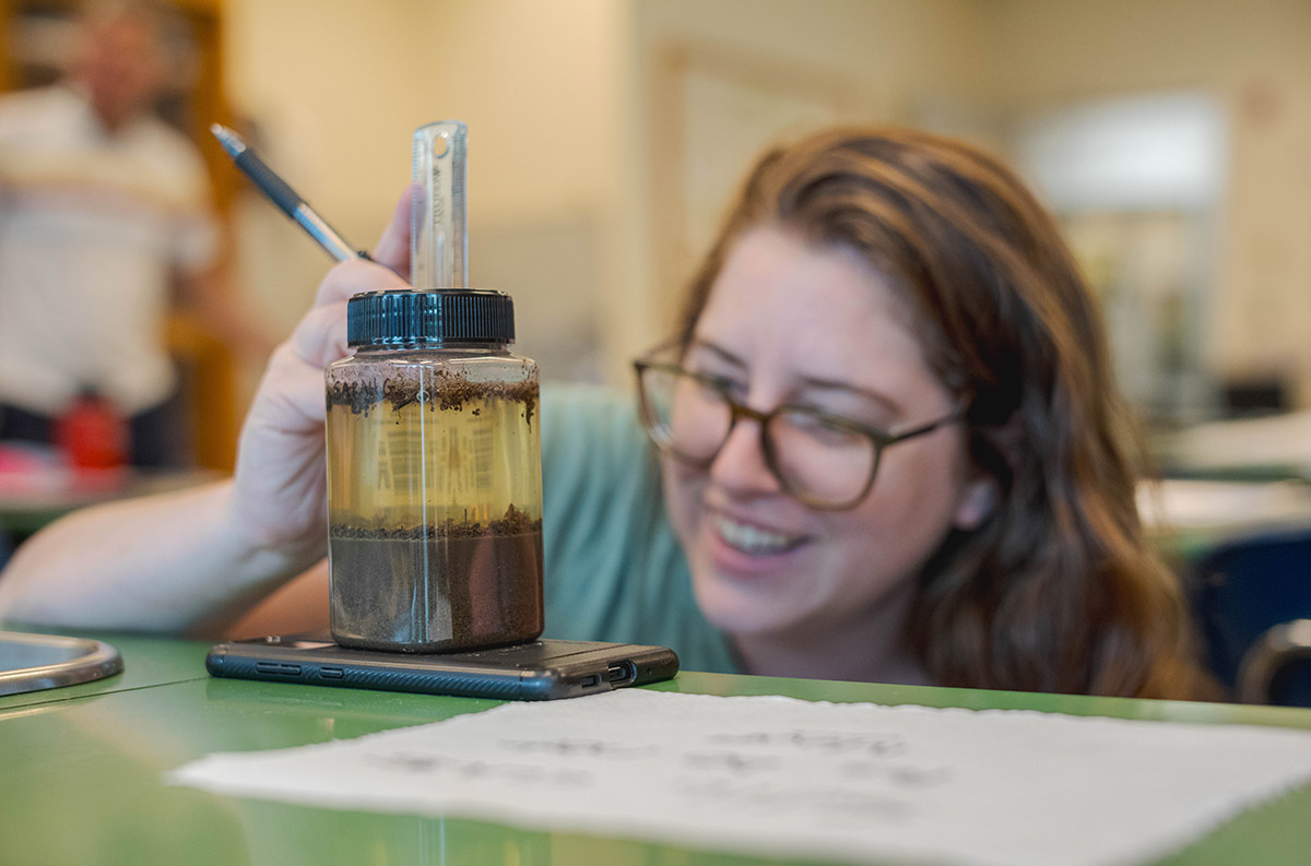 A person with long brown hair, wearing glasses, observes plant and liquid matter in a jar