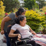 A person pushes another person in a wheelchair through a sunny garden setting