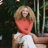 a headshot of a woman with curly blonde hair, an orange long sleeved top and white pants, sitting on a stool and surrounded by plants