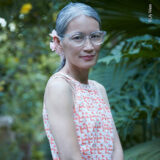 A person in an orange and white shirt and translucent glasses poses for a photo among green foliage