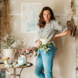 A person in blue jeans poses for a photo in a rustic room full of flower arrangements