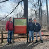 Three people gather around a garden sign for a photo on a winter day