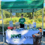 A person wearing a face mask and a green T-shirt waits behind a booth table to help with composting questions