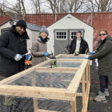 Four people in winter jackets and wool caps use hammers to secure a new three-bin compost system