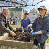 Three people in sweatshirts and hats pose for the camera as they work with compost in a wooden bin