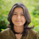 A person in a brown shirt with long dark hair poses outdoors for a photo