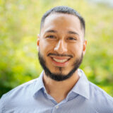 A person in a blue button up shirt with a dark beard poses outdoors for a photo