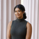 A person in a black turtleneck with dark hair tied back poses for a photo in front of a pair of stone pillars