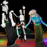 three giant skeleton puppets in costumes dancing