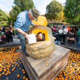 a person inside of a giant pumpkin with another person standing next to the pumpkin with a white bucket