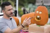 a person with dark brown hair holds a paintbrush in front of a carved pumpkin with large white eyes and two front teeth