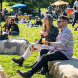 two people sitting on bales of hay in a field enjoying glasses of beer in the sunlight