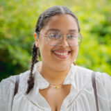 A person in a white shirt with dark braided hair and glasses poses for a photo outdoors