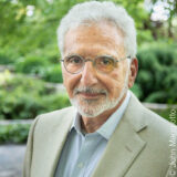 A person with short white hair, wearing glasses, a blue dress shirt, and a tan blazer, poses outdoors for a photo