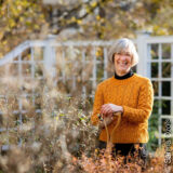 A person with short gray hair, wearing an orange cable-knit sweater, poses with a shovel in their fall garden