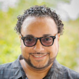 A person in a black shirt with tinted glasses and a dark beard poses outdoors for a photo