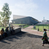 A view of a geometric building on a sunny day, with people walking and sitting among the paths and benches of this curated space