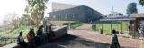 A view of a geometric building on a sunny day, with people walking and sitting among the paths and benches of this curated space