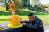 a person sitting down in a leather jacket carving a pumpkin with a monkey face and smaller pumpkins on top of it