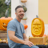 A person in a gray shirt with short dark hair poses for a photo in front of a pumpkin face he's just carved into an orange gourd