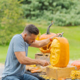 Brown hair man leaning over wearing a grey shirt holding a tool with a sharp edge pointed into an orange pumpkin face that is being carved to look like a human face. A carved out jack-o-laterned with a cut up mouth is in the background of the sculptural looking carved face of the main pumpkin.