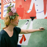 A person with blonde hair, wearing a crown of flowers, creates art on a paper-covered wall