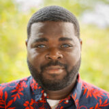 A person in a red and blue shirt with a dark beard poses outdoors for a photo