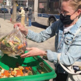 A person in a black face mask and blue denim jacket drops a bag of food scraps into a green bin