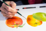 A hand uses a paint brush to paint realistic apples
